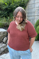 Woman standing in a backyard patio with greenery in the background. She wears a reddish brown crochet t-shirt and pale blue pants. The tee has an all-over texture.  thumbnail