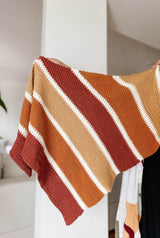 Person offscreen holds a crochet shawl in a white room. The shawl is striped in brick red, saffron orange, pale peach, and dark wine red. Each stripe is separated by a smaller stripe of cream.  thumbnail