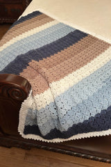 Crochet striped baby blanket laid on bed over white quilt. The blanket is made in shades of blue, white, and brown, with a lacy white border.  thumbnail