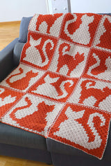 Orange and cream crochet blanket draped over a charcoal grey couch. The blanket is made in individual blocks featuring four different cat poses.  thumbnail