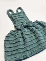 Flat lay of knit pinafore dress on white background. The skirt and bodice have a ridged texture and a bow at the waist.  thumbnail