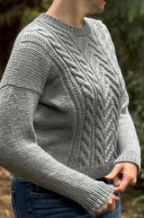 Side view of woman wearing grey sweater with cables on the front and plain sleeves, sides, and back. The sweater has long sleeves and a drop shoulder construction. thumbnail