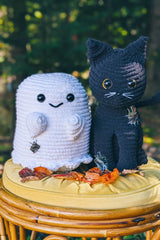 Crocheted ghost and black cat sit on a yellow stool in front of trees. They both have sewn-on patches, plastic eyes, and embroidered features. thumbnail