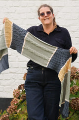 Woman in black top and dark jeans stands in front of a white painted wall and hydrangea bush. She holds up a knit shawl or scarf that is color blocked in denim blue, sage green, and golden yellow, with each block separated by cream. thumbnail