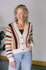 Woman standing in front of a plain wall with a potted plant in the background. She wears a crochet cardigan with vertical stripes over a brown tank top and jeans. thumbnail