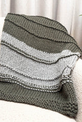 Knit blanket draped over cream armchair, in front of a cream drapery. The blanket is color-blocked in grey-green and pale grey. thumbnail