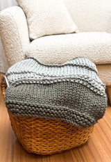 Knit blanket folded in wicker basket, sitting in front of cream armchair. thumbnail