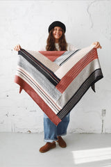 Woman in a black hat, white sweater, and jeans standing in an all-white room. She holds a knit shawl toward the camera. The shawl is made of blocks of different textures in terracotta, cream, pale blue, beige, and black. thumbnail