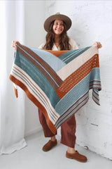 Woman standing in an all-white room holding a shawl toward the camera. She wears a flat brimmed hat, cream shirt, and rust red pants. The crochet shawl is color blocked in rusty orange,  dark sage green, cream, chocolate brown, and pale blue. thumbnail
