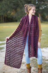 Woman standing in an open grassy area with trees behind. She wears a large lacy crochet stole in a dark red-purple color, over a tan top and light colored jeans. thumbnail