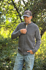 Man standing in a wooded area, wearing a zippered crochet cardigan, baseball hat, and jeans. thumbnail
