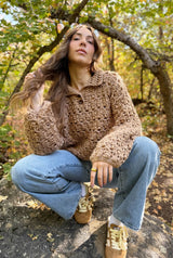 Woman crouched on boulder in early fall woods. She wears sneakers, jeans, and a crochet pullover with long sleeves and a polo collar. thumbnail