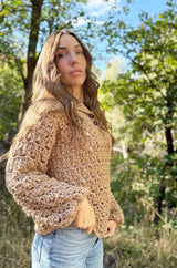 Side front view of woman wearing crochet pullover. It has long sleeves with a blousy cuff, button placket at top neck, and a lacy texture. The sweater is a warm tan beige. thumbnail