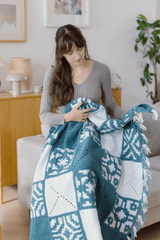 Woman sitting in a pale living room, on the arm of a couch. She has a crocheted afghan on her lap, made of multiple square patterns. It has fringe along the edge. thumbnail