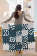 Woman standing in a pale room holds a folded crochet blanket. The blanket is made in white and medium blue, with solid squares in those colors and patterned squares mixing those colors. It had mixed fringe on the shorter top and bottom edges. thumbnail
