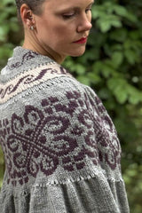Side view of woman wearing knit shawl, looking over her right shoulder. The shawl is knit in pale grey with bands of colorwork in dark purple and pale peach. thumbnail