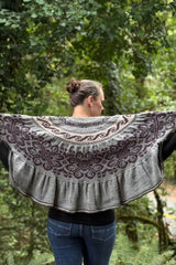 Back view of woman holding semi-circular shawl open. The top edge of the shawl is straight, and there are bands of filigree patterns on a grey background. The colorwork is knit in dark purple and pale peach. thumbnail