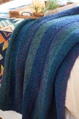 Knit blanket draped over the edge of a bed, with a wooden tray in the background. The blanket is knit in a color changing yarn in shades of blue, green, and purple. The throw also has horizontal textured stripes of stockinette and garter. thumbnail