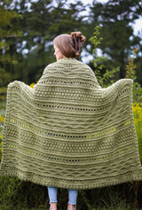 Woman with back to camera holds a crochet aghan across her back. It is a pale green with bands of textured patterns. thumbnail