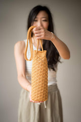 Woman holds a crochet water bottle holder toward camera, standing against a beige backdrop. thumbnail