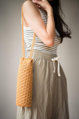 Side view of woman wearing water bottle holder over shoulder, with long strap. The top of the holder is at her hip. The bag is made with a solid textured fabric, in a warm camel color. thumbnail