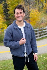 Man in a white shirt and dark pants stands outside with fall foliage and a white wooden fence behind. He also wears a knit jacket with a collar, button band, and pockets. thumbnail
