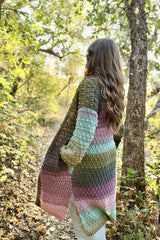 Side back view of woman standing in the woods. She wears a long crochet cardigan, with long sleeves, pockets, and a split hem. thumbnail