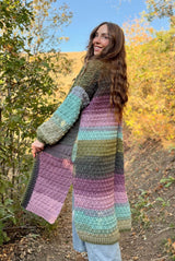 Side back view of woman in long crochet cardigan with her face turned toward camera. The cardigan is made in self-striping yarn in shades of green, blue, pink, and purple. The cardigan hangs to mid-calf and has a split hem up to the knee. The sleeves are long and there are also side pockets. thumbnail