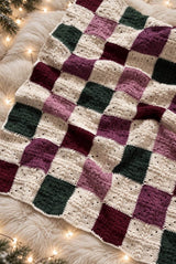 Crochet patchwork blanket laid on a white faux-fur rug. The blanket squares are cream, dark green, burgundy, dark pink, and medium pink. thumbnail