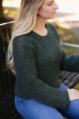 Side view of woman wearing green crochet pullover with long sleeves, sitting on a bench. thumbnail