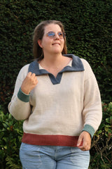 Woman in a knit polo sweater standing in front of a green hedge. The sweater is mainly cream, with a terracotta hem, sage green cuffs, and a denim blue collar. thumbnail